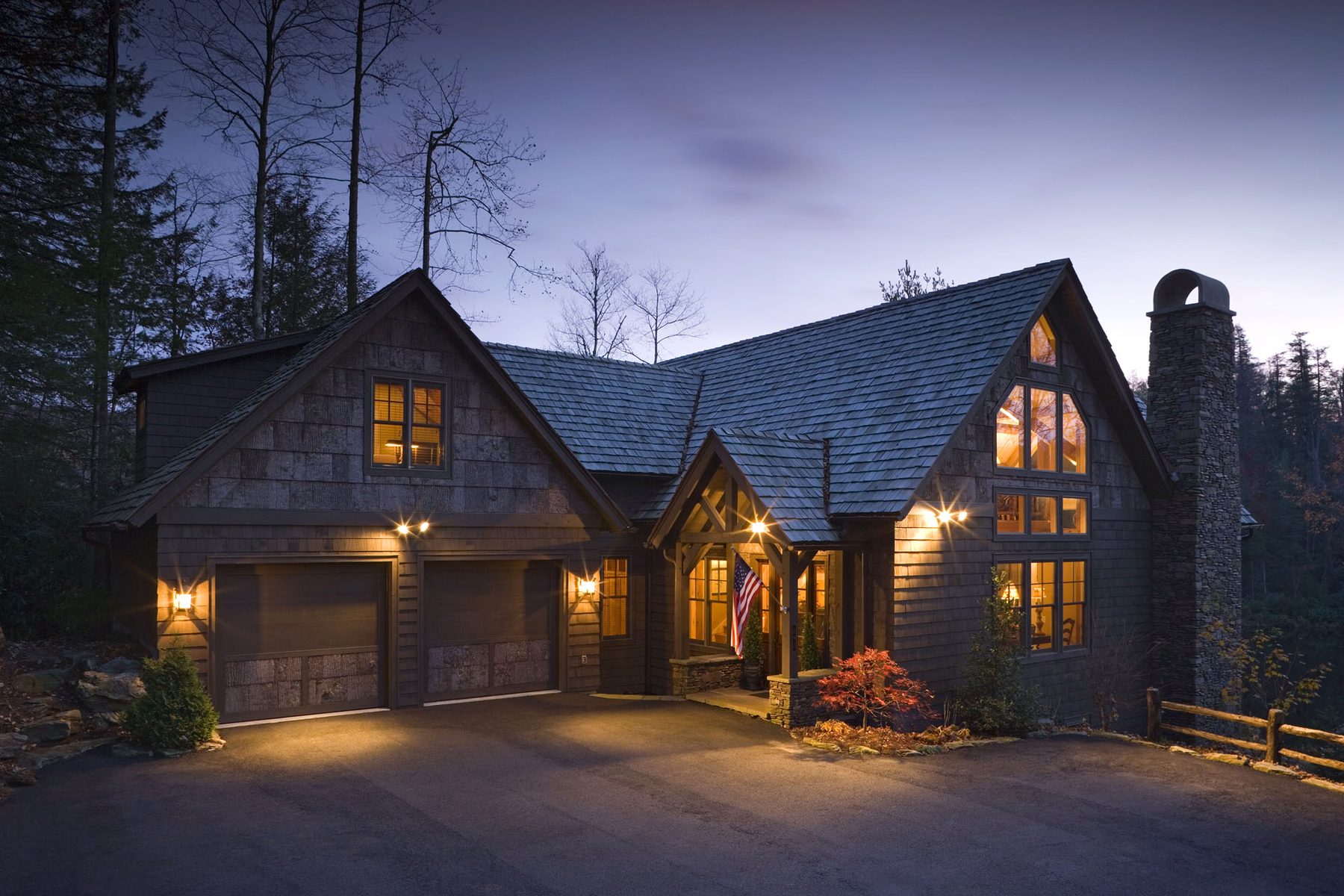 Entry elevation at dusk — lantern-lit gables, cedar shingles, and the dry-stack stone chimney